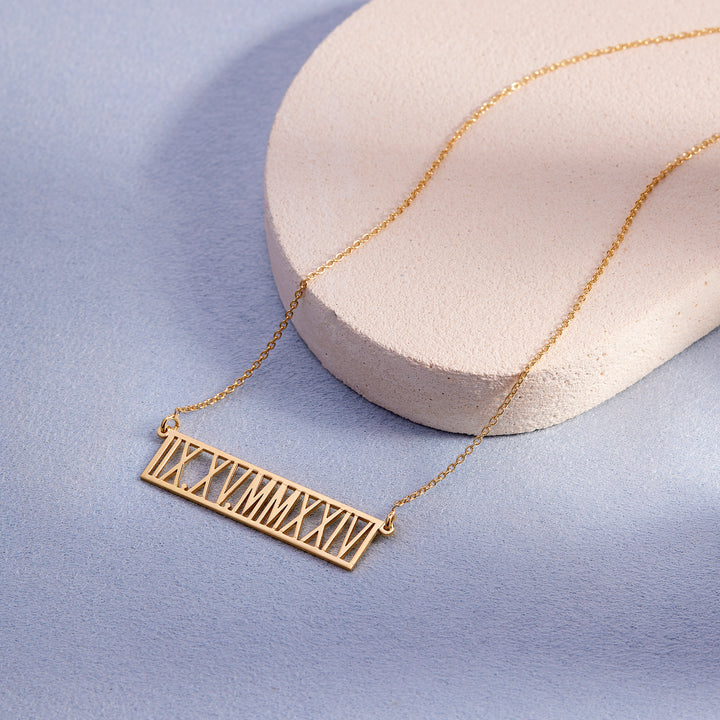 Gold necklace with Roman numeral pendant on a light blue background