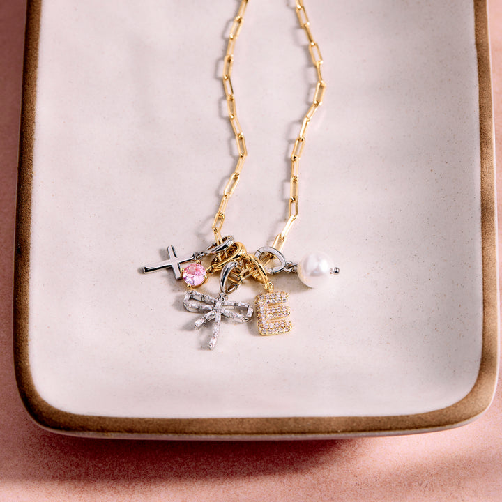 A gold chain necklace with multiple charms, including a pink gemstone cross, a silver bow, a gold letter &