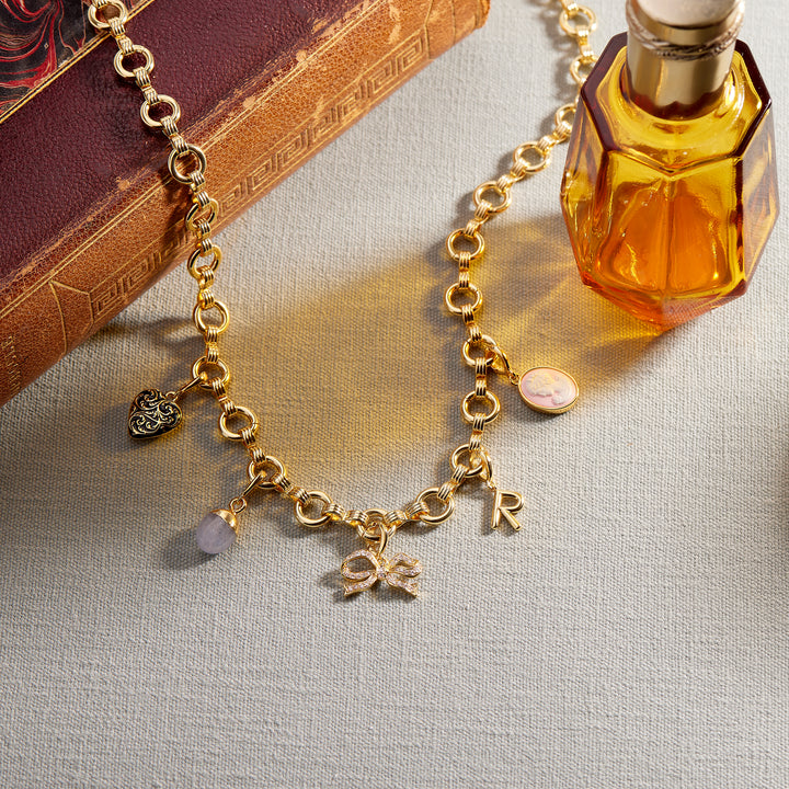 A gold-tone charm necklace with multiple dangling charms, including a heart, a small oval charm, a bow, and letters, displayed on a textured beige fabric next to a vintage book and an amber glass bottle.