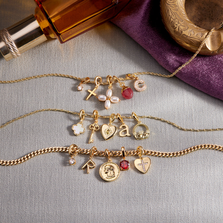 Three gold-toned chain necklaces with assorted religious and decorative charms are arranged on a light gray fabric surface. The necklaces feature charms including a cross, flower, pearl-like beads, pink gemstone, letter &