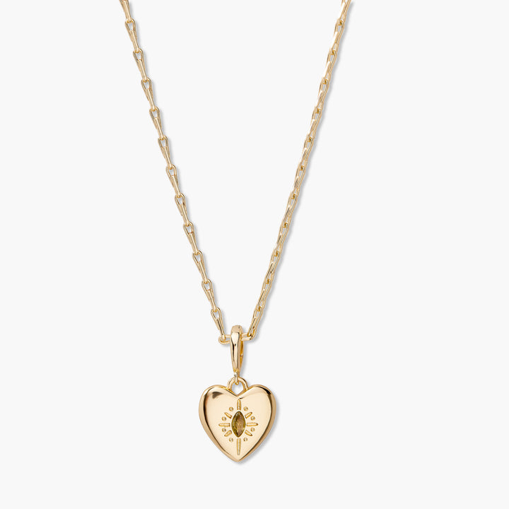 A gold heart pendant necklace with a sunburst design and a central marquise-cut gemstone, suspended from a delicate gold chain against a light off-white background.