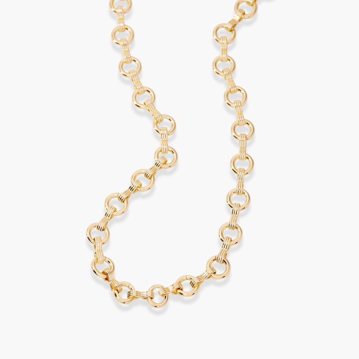 A gold chain necklace with textured, ribbed oval links, displayed on a clean, light off-white background.