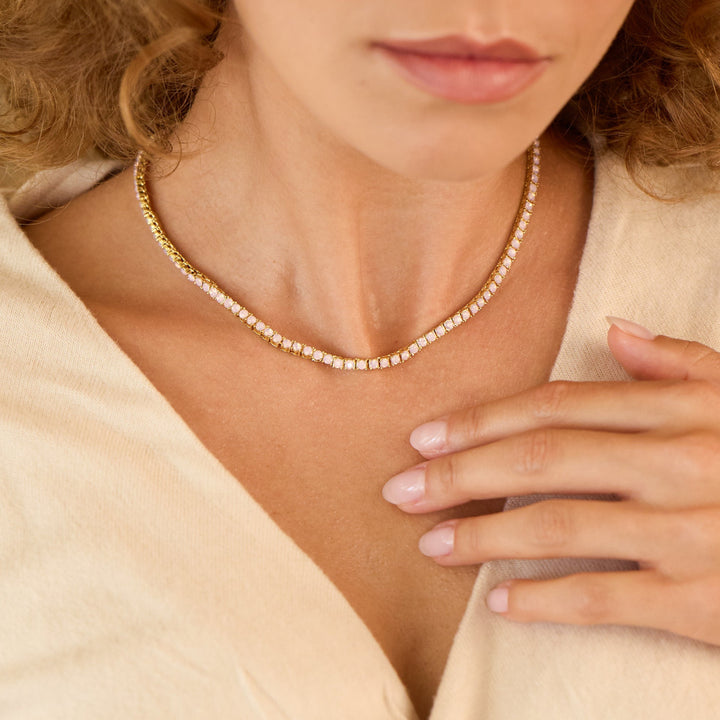 Woman wearing a beige top with a knot detail, pearl necklace, and hand on chest.
